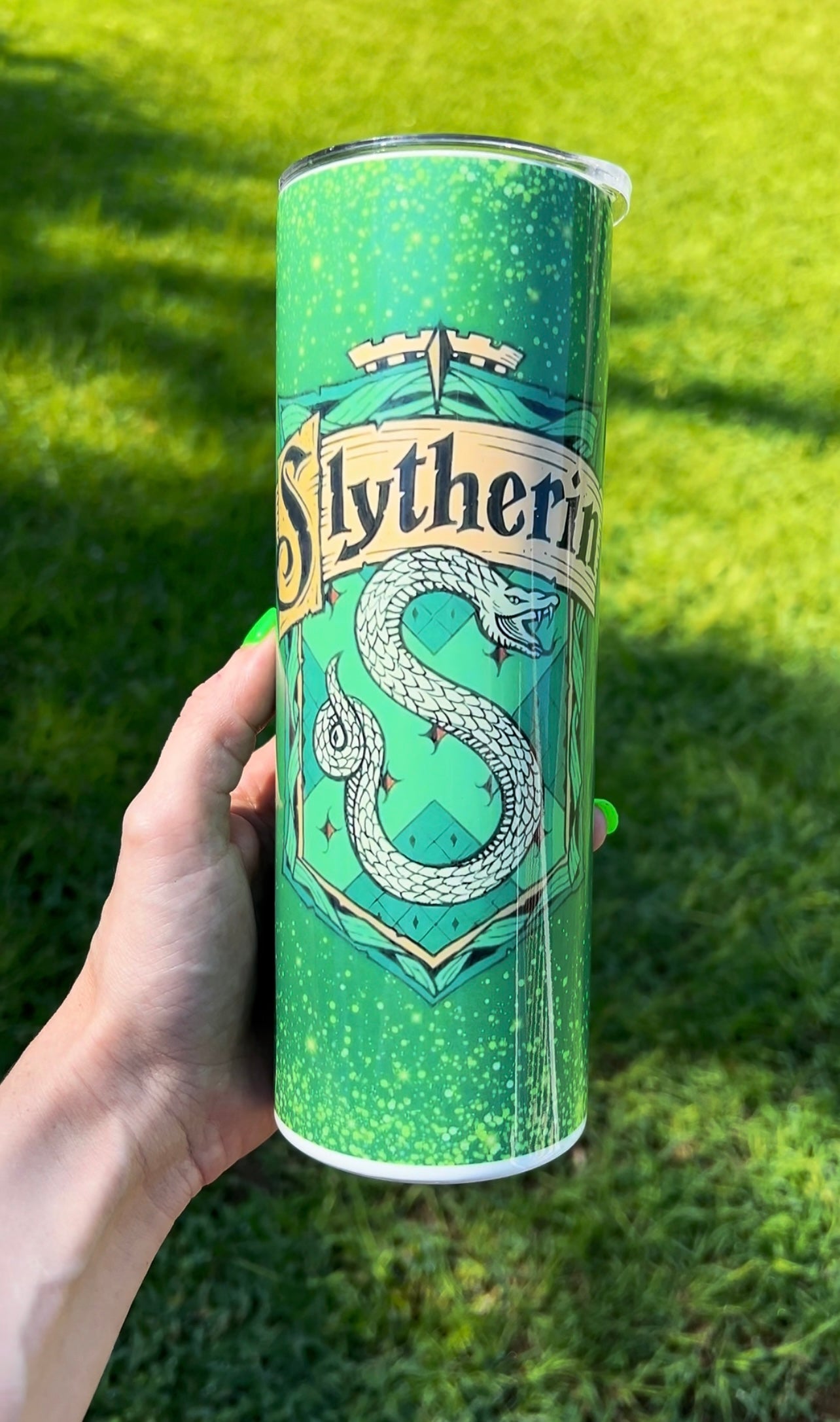 Handheld 20oz skinny tumbler featuring the Slytherin house crest and silver serpent over a sparkling green emerald galaxy background with a grassy outdoor backdrop.