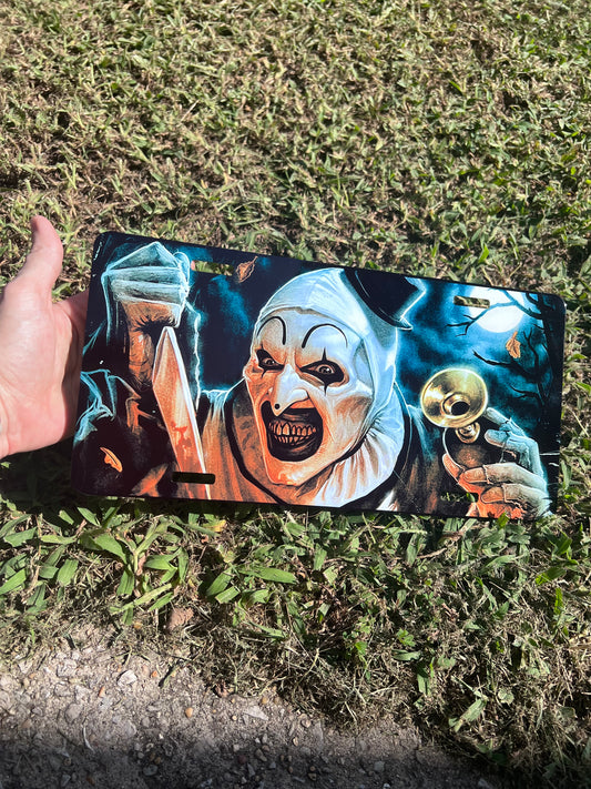 Art the Clown Terrifier novelty front license plate resting flat, showcasing the creepy clown with a bloody blade and horn under a moonlit sky.