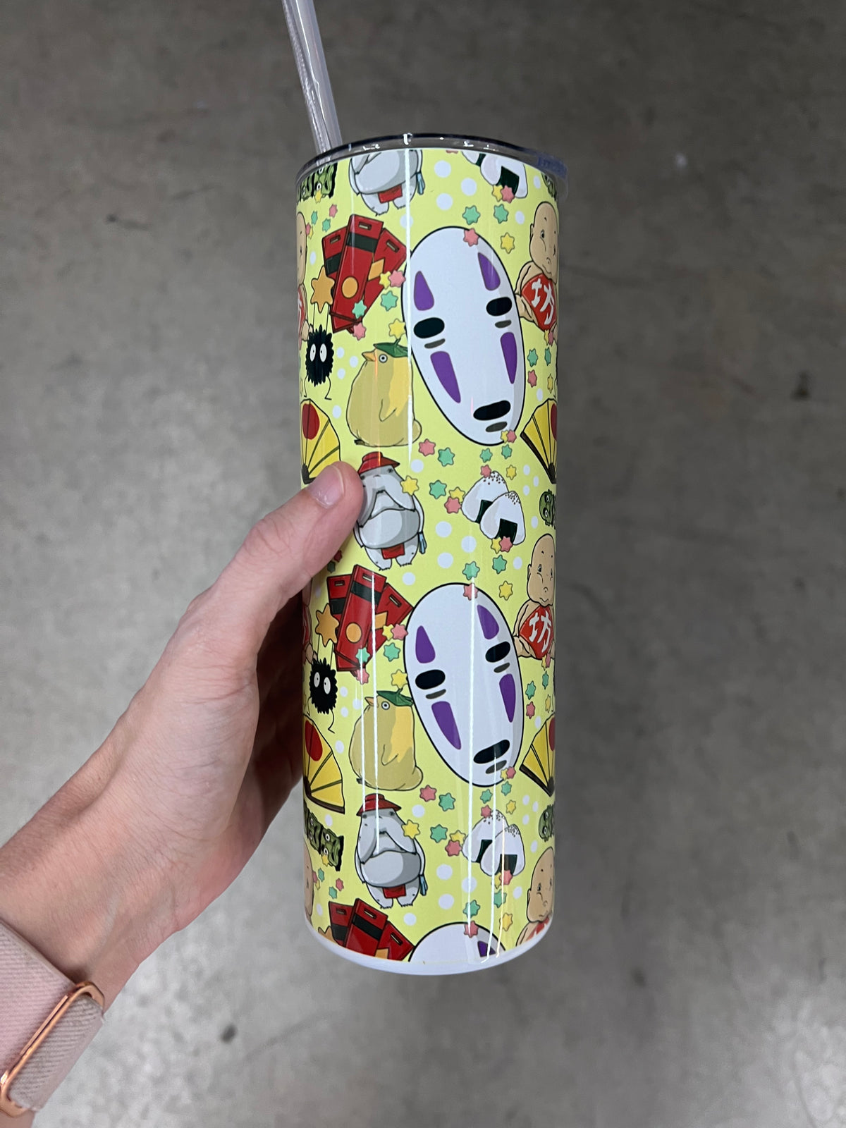 A hand holding a custom pastel yellow 20oz tumbler featuring a repeating Studio Ghibli pattern with No-Face, soot sprites, and Boh.