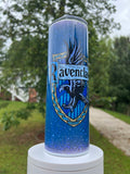 20oz skinny tumbler featuring the Ravenclaw house crest with an eagle over a blue galaxy and starry glitter background, displayed on a white stand outdoors.