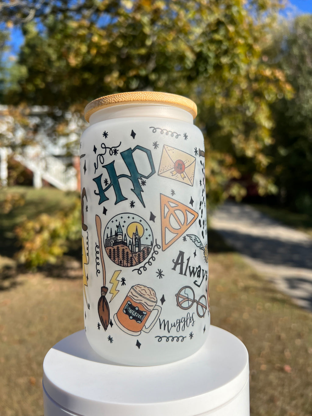 16oz frosted glass can tumbler with a bamboo lid, decorated with a seamless pattern of magical wizarding world icons like the Sorting Hat, flying broomsticks, lightning bolts, and butterbeer, displayed outdoors on a white stand.