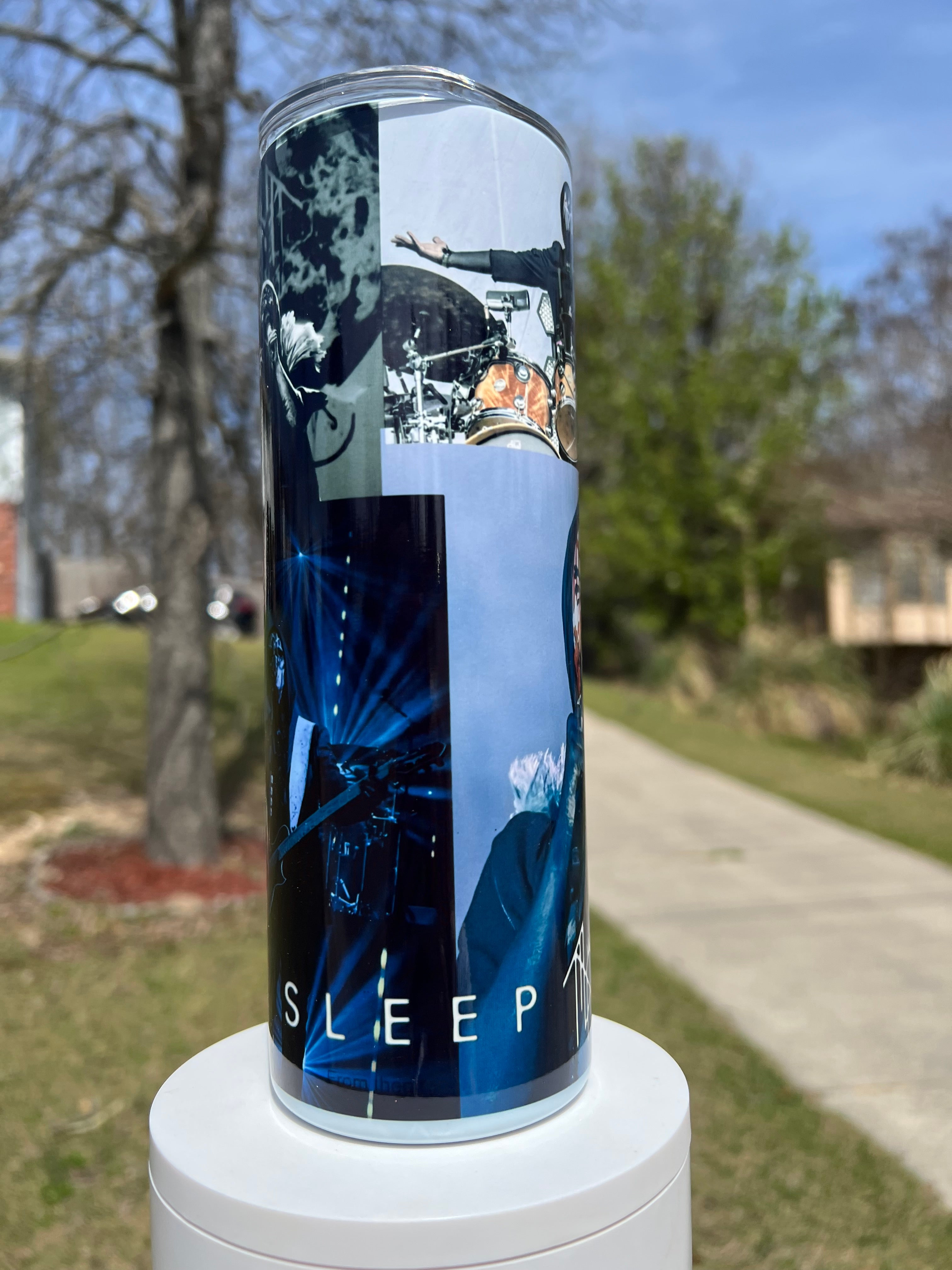 A close-up view of a 20oz skinny tumbler featuring Sleep Token band members. At the top, a figure plays a detailed drum kit; at the bottom, Vessel sings into a microphone with his iconic red face paint. The bottom edge has black band logos.