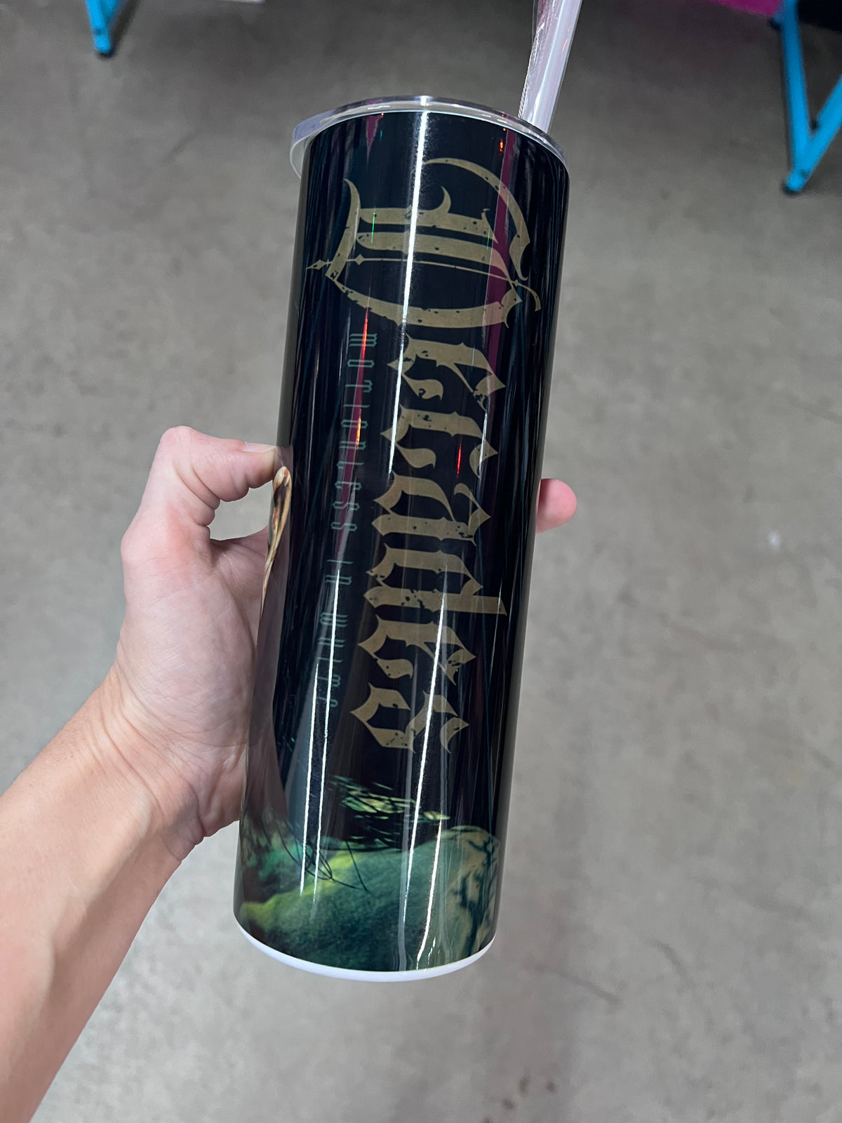 A hand holding a custom black tumbler featuring the word "Decades" in large, gold gothic lettering with "Motionless In White" printed vertically next to it, fading into an eerie green bottom.