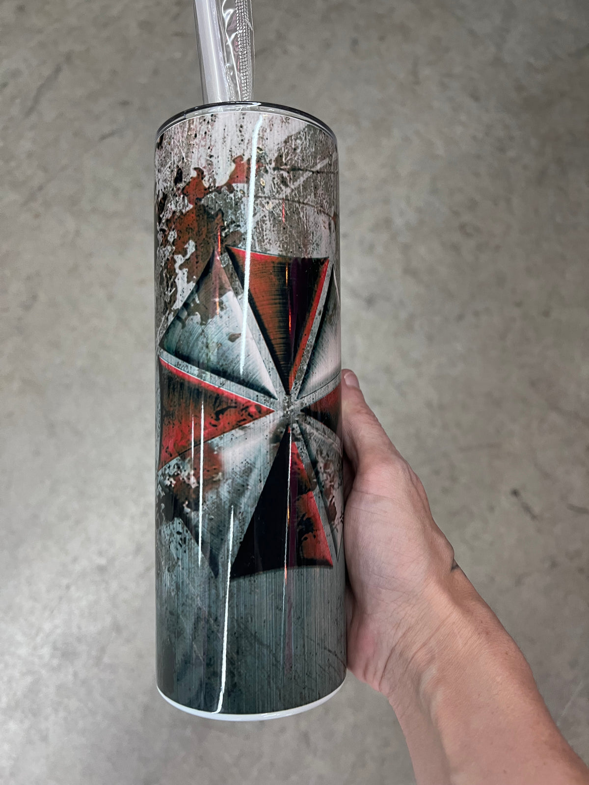 A hand holding a custom tumbler featuring the red and white Umbrella Corporation logo set against a heavily distressed, scratched concrete and metal background.