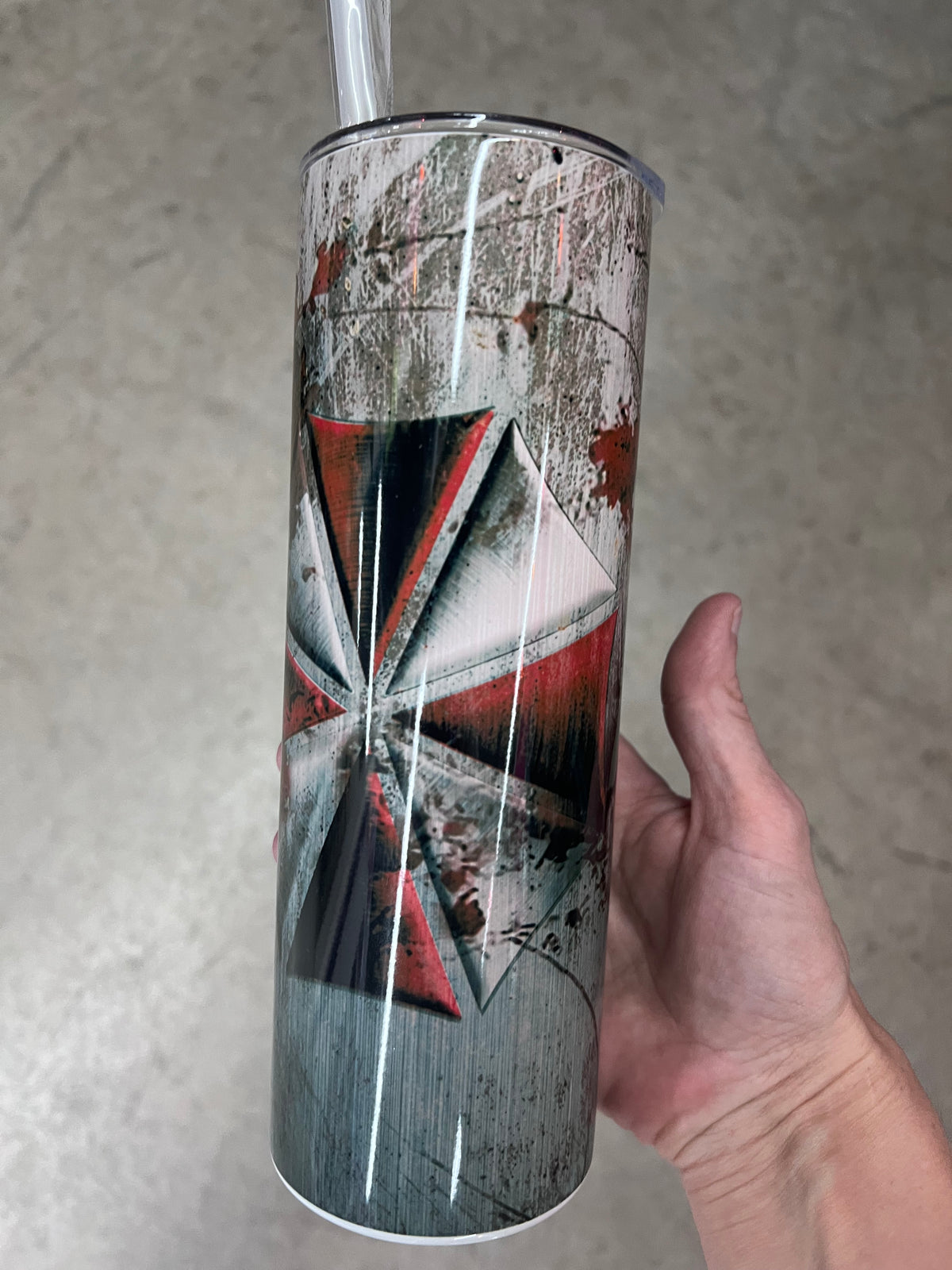 A hand holding a custom tumbler featuring the red and white Umbrella Corporation logo set against a heavily distressed, scratched concrete and metal background.